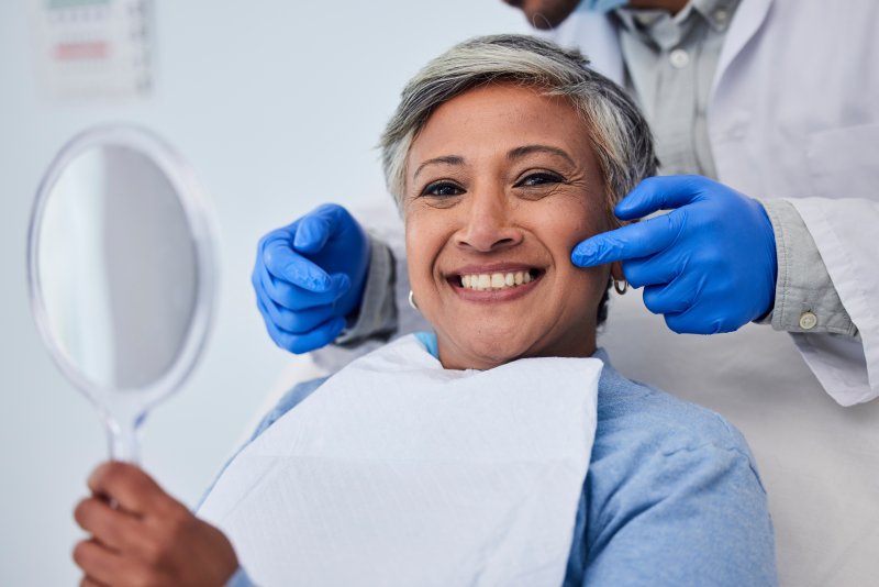 Older woman smiling with teeth at viewer while holding handheld mirror in front of her as dentist stands behind and points gloved fingers at each side of her face